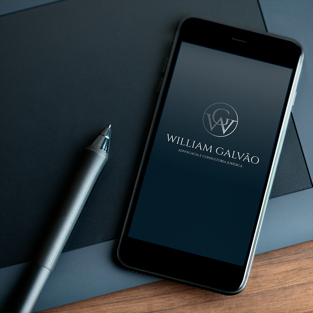 Smartphone sobre mesa de madeira escura, exibindo a identidade visual da marca “William Galvão – Advocacia e Consultoria Jurídica” com logotipo elegante nas iniciais WG em cinza sobre fundo azul-escuro. Ao lado, uma caneta moderna prateada. Imagem de estilo corporativo e minimalista, transmitindo credibilidade e sofisticação.